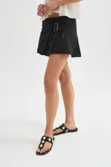 Minifalda de jean negra con diseño cruzado, cierre de hebilla metálica y detalle de tablones plisados en el lateral.