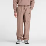 Pantalón deportivo New Balance modelo Woven Pant, color marrón topo, de tejido ligero y corte ancho. Presenta cintura elástica con cordón ajustable y un pequeño logo New Balance bordado en la pierna izquierda.