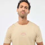Remera beige de manga corta y cuello redondo, con pequeño estampado circular en el centro del pecho que incluye texto en tonos rosados y blancos.