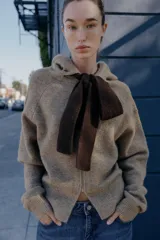 Chaqueta de punto con capucha, manga larga y cierre frontal con cremallera. Presenta un diseño en color beige con un lazo decorativo a contraste en color marrón oscuro en la zona del cuello.