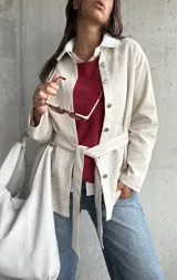 Sobrecamisa de gamuza en color beige, con cierre frontal de botones metálicos, cuello camisero y lazo ajustable en la cintura. Presenta un diseño de corte recto y mangas largas.