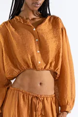 Camisa crop de manga larga confeccionada en crepé color tostado, con cierre frontal de botones y elástico en la cintura.