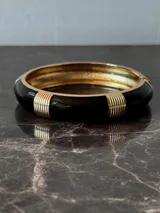Pulsera rígida tipo brazalete, color negro con detalles dorados.