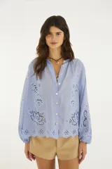 Blusa celeste de algodón con bordado calado y ojales en el delantero, mangas amplias y ruedo. Presenta escote redondo con cierre frontal de botones.