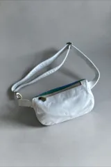 Riñonera blanca con solapa de cuero tornasolado, cierre frontal superior y correa ajustable. Presenta detalles de arte pintado a mano en el frente con inscripciones y manchas abstractas en tonos verdes, marrones y negros.