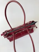 Cartera de cuero de tamaño medio con estructura rígida. Presenta una tira decorativa con tachas y hebillas metálicas en los extremos, además de lazos laterales. Cuenta con doble asa para llevar al hombro y cierre superior.