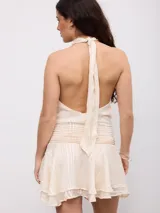 Vestido corto color beige con diseño a rayas sutiles, cuello halter y espalda descubierta. Cuenta con una faja fruncida en la cintura y falda con volados en el borde inferior.
