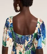 Vestido largo de viscosa con estampado tropical de flores blancas y hojas verdes y azules. Presenta escote cuadrado con lazo ajustable en el centro del busto y mangas cortas abullonadas. La falda es escalonada con volados.