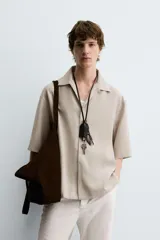 Camisa de corte relajado color beige, con cuello bowling, manga corta y cierre frontal con botonadura oculta por solapa. Presenta aberturas laterales en el bajo.