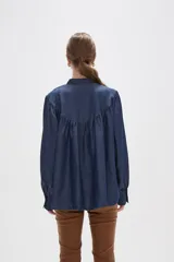 Blusa de lyocell en tono azul denim, con diseño holgado y frunces en el canesú. Presenta cuello alto con volados, cierre frontal mediante cordón ajustable y mangas largas con puños abotonados.