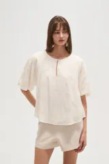 Blusa blanca de manga corta con escote redondo y abertura frontal con botón. Presenta bordados florales en tono beige distribuidos por toda la prenda.