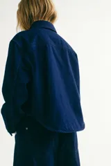 Camisa de denim rígido de algodón azul oscuro, con corte relajado, hombros caídos, cuello clásico, botones ocultos a presión, bolsillos superiores con solapa y bajo redondeado.