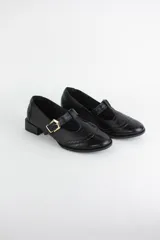 Zapato tipo guillermina estilo oxford, confeccionado en cuero negro con detalles de perforaciones decorativas en la puntera y cierre mediante tira con hebilla metálica dorada. Presenta un taco bajo cuadrado.