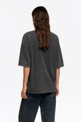 Remera de algodón, color gris oscuro con efecto gastado (acid wash), corte oversize y escote redondo. Presenta un estampado frontal artístico con una ciudad, flores amarillas y una bota de lluvia verde.