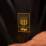 Camiseta de fútbol Puma del Club Atlético Peñarol, edición aniversario, modelo de manga larga. Diseño con mitad derecha a rayas verticales amarillas y negras, y mitad izquierda negra con escudo del CURCC y logo de Puma en dorado. Presenta cuello polo negro y sponsor antel en el pecho.