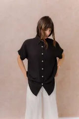 Camisa negra de calce amplio y relajado, confeccionada en tejido liviano con textura sutil. Cuenta con diseño oversize, hombros caídos, mangas cortas amplias, cuello camisero y abotonadura frontal con botones claros.