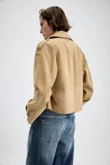 Trench corto cruzado de piel de ante color tostado, con cuello de solapa, bolsillos delanteros de vivo y manga larga con trabillas en los puños.