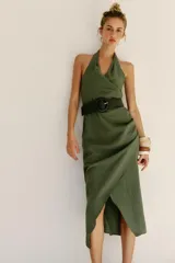 Vestido midi verde militar de cuello halter con escote en V cruzado y espalda descubierta. Cuenta con pliegues laterales, bajo asimétrico y un cinturón incorporado con hebilla redonda oscura.