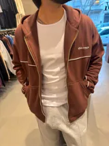 Campera con capucha de corte boxy color marrón, con cierre frontal completo y bolsillos tipo canguro. Presenta una línea blanca horizontal en el pecho que se extiende por las mangas y un pequeño bordado con texto en el pecho.
