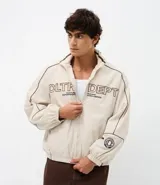 Campera de tejido técnico color beige con detalles de costuras en contraste color marrón. Presenta cuello alto, cierre frontal con cremallera, mangas largas con puños elásticos y estampa frontal con texto. Incluye un detalle bordado en la manga.