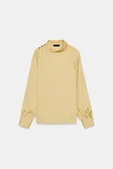 Blusa de manga larga color amarillo pálido, con cuello alto drapeado y detalle de botones dorados en el hombro izquierdo. Las mangas terminan en puños anchos.