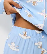 Pijama largo estilo americano de poliéster, compuesto por una camisa de manga larga con cuello solapa y cierre frontal de botones, y un pantalón largo de corte recto. Presenta un estampado integral del personaje Cinnamoroll sobre un fondo celeste.