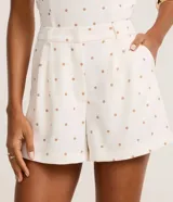 Bermuda de piquet color blanco con estampado de lunares negros, de corte recto, tiro alto, con trabillas en la cintura, pliegues y cierre tradicional.