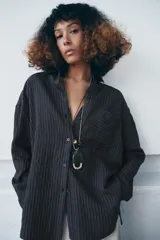 Camisa fluida de corte oversize confeccionada en mezcla de viscosa, con diseño de rayas verticales finas en tono beige sobre fondo blanco. Presenta cuello solapa, cierre frontal con botones, manga larga con puños y un bolsillo de plastrón en el pecho.