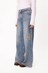 Jean de corte wide leg en denim azul claro con efecto lavado. Presenta bordados del logo de la marca en color ocre en ambos laterales a la altura de las rodillas y ruedo desflecado.
