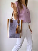 Bolso tipo tote de cuero color lila con textura croco, detalles en cuero marrón y laterales con franjas blancas. Incluye manijas de cuero marrón y accesorio de borlas colgantes.