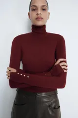 Jersey de punto liso en color beige, con cuello alto y manga larga con detalle de botones metálicos en los puños.