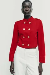Cazadora de estilo militar en color rojo, con cuello redondo, manga larga y cierre frontal mediante alamares con botones dorados. Presenta trabillas con botón en los hombros y detalle de costuras en los puños.
