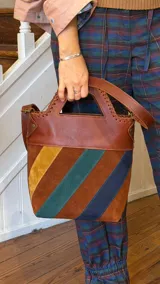 Bolso tipo matera confeccionado en cuero vacuno, con diseño estructurado y manija superior integrada. Presenta un cuerpo con franjas diagonales en gamuza de colores contrastantes (amarillo, verde y azul) sobre una base de cuero marrón, con detalles de costuras artesanales en los bordes.