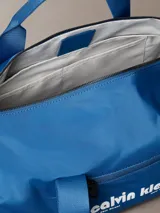 Cartera tipo duffle deportiva de color azul brillante, confeccionada en poliéster. Presenta asas de mano y una correa ajustable para el hombro. En el centro tiene estampado el logo de Calvin Klein en blanco, incluyendo la dirección "West 39th Street New York City". Posee un bolsillo exterior con cierre en el frente.