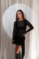 Vestido corto negro de encaje transparente, con mangas largas y falda ajustada con detalle lateral asimétrico tipo pareo.