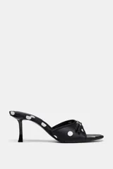 Sandalia tipo mule negra con estampado de lunares blancos, efecto satinado, punta fina, tacón fino y tira delantera con lazo decorativo.