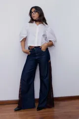 Pantalón de jean estilo wide leg, confeccionado en denim rígido de color azul oscuro. Presenta paneles laterales de encaje con diseño floral en tono rojizo y terminaciones deshilachadas en el ruedo.