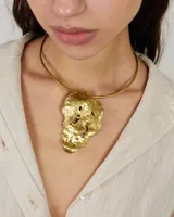 Gargantilla rígida de metal dorado con dije central de diseño orgánico y textura irregular. El diseño permite múltiples usos, incluyendo la opción de utilizar la gargantilla sola o con el dije.