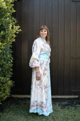 Vestido largo estampado con motivos florales en tonos pastel y detalles de cadenas doradas. Presenta cuello mao, cierre frontal con botones variados, mangas largas con puños elásticos y un cinturón a tono con hebilla dorada que realza la cintura. El ruedo del vestido tiene una franja de color celeste.