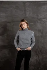 Buzo de punto acanalado color gris, con cuello alto y cierre frontal parcial. Presenta detalles de costuras decorativas con patrón de bloques en color blanco en los hombros, puños y ruedo.
