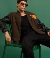 Campera bomber de estilo college con cuerpo a cuadros en tonos marrones y mangas largas de cuero sintético negro. Presenta cuello, puños y cintura acanalados a rayas, cierre abotonado, bolsillos laterales y aplique bordado de letra "B" en el pecho.