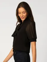 Blusa de color blanco con escote en V y detalle de lazo frontal. Presenta mangas cortas de gasa con terminación en puño.