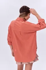 Camisa color coral de corte holgado, con cuello clásico, cierre frontal con botones, mangas largas con puños abotonados, dos bolsillos de parche en el pecho y ruedo asimétrico.
