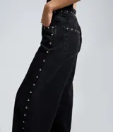 Pantalón wide leg de denim negro con acabado desgastado, tiro medio y aplicación de tachas metálicas a lo largo de los laterales.