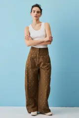 Pantalón marrón de corte ancho, tiro alto y diseño holgado, adornado con pequeños apliques metálicos distribuidos por toda la prenda.