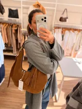 Cartera tipo hobo de textura gamuzada en color marrón oscuro, con diseño flexible y asa larga regulable. Presenta un detalle de flecos largos desmontables en uno de los laterales y cierre superior.