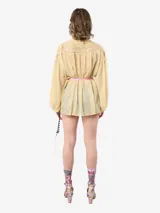 Blusa de gasa color beige con mangas abullonadas, cuello mao y cierre frontal con botones. Presenta aplicaciones de flores bordadas con lentejuelas en el canesú delantero y un lazo de satén rosa en la cintura.
