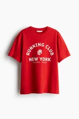 Remera de algodón color rojo, con cuello redondo y manga corta. Presenta un estampado frontal en blanco con el texto "Running Club New York Central Park - Manhattan" y un logo circular con una 'R' en el centro.