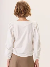 Camisa blanca con mangas largas abullonadas que terminan en puño elástico y detalle de volados en los hombros y puños. La espalda es de tejido de punto.