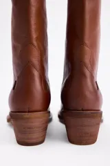 Bota estilo western de caña media, confeccionada en gamuza color marrón con bordados decorativos en hilo claro. Cuenta con taco cuadrado de madera y tiradores laterales.